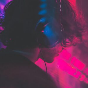 A person with long hair, likely a musician, wears headphones in the MOON MUTT Recording Studio, surrounded by colorful lights that create a vibrant atmosphere pulsating with music.